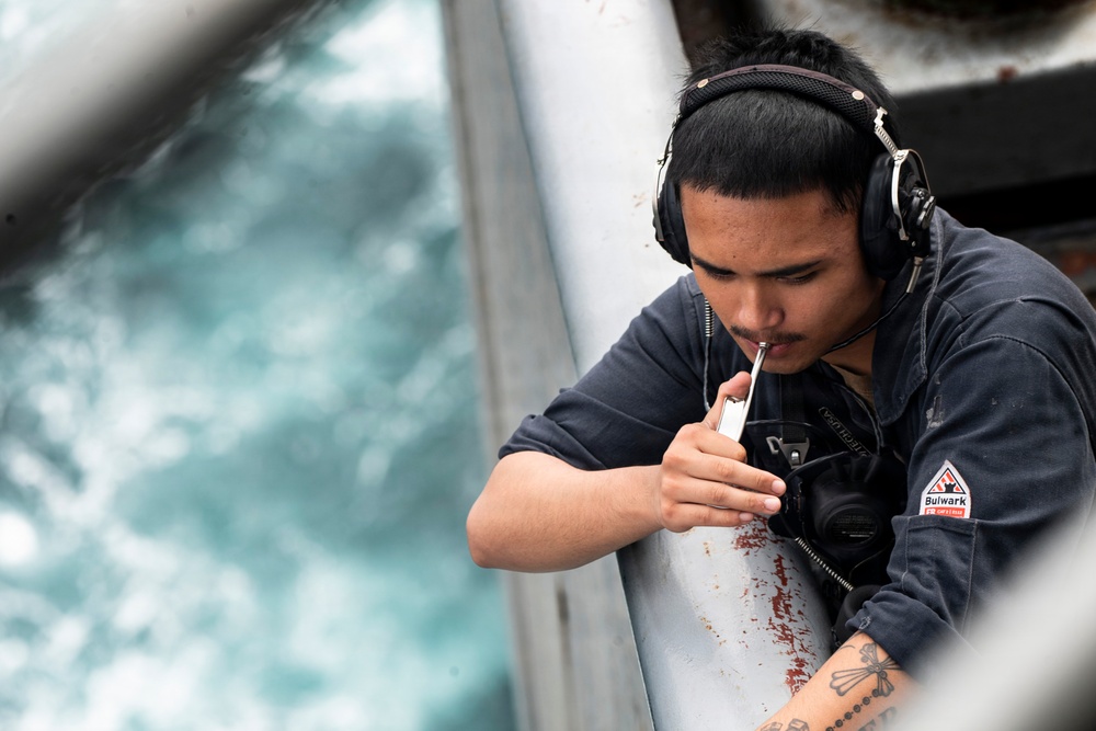 A young man wearing communication headphones and a dark navy 'Bulwark FR' flame-resistant work shirt is focused on a task involving a small tool or instrument near a metal structure. He appears to be working on or inspecting a component on what looks like a vessel or industrial platform, with churning water visible in the background. A cross tattoo is visible on his forearm.