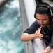 A young man wearing communication headphones and a dark navy 'Bulwark FR' flame-resistant work shirt is focused on a task involving a small tool or instrument near a metal structure. He appears to be working on or inspecting a component on what looks like a vessel or industrial platform, with churning water visible in the background. A cross tattoo is visible on his forearm.