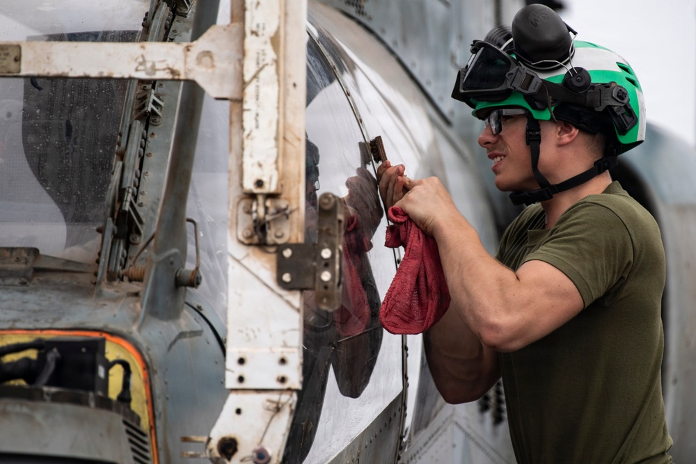 A military aviation technician or deck crew member is performing maintenance on an aircraft, likely a helicopter, using a tool and a red shop rag. The individual is wearing a green flight deck helmet with goggles and a green t-shirt, consistent with U.S. Navy flight deck crew attire. The aircraft surface shows signs of wear, corrosion, and weathering typical of carrier-based or maritime operations.
