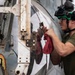 A military aviation technician or deck crew member is performing maintenance on an aircraft, likely a helicopter, using a tool and a red shop rag. The individual is wearing a green flight deck helmet with goggles and a green t-shirt, consistent with U.S. Navy flight deck crew attire. The aircraft surface shows signs of wear, corrosion, and weathering typical of carrier-based or maritime operations.