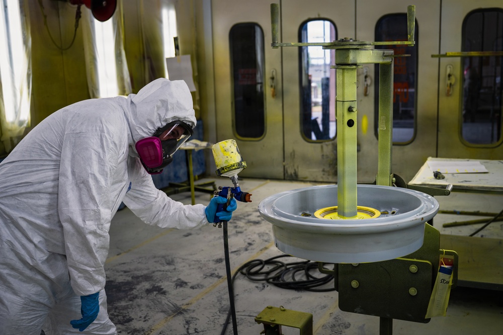 A worker in full protective gear is spray painting a large circular component, likely a vehicle wheel or rim, mounted on a rotating stand inside an industrial paint booth. The worker is wearing a white Tyvek suit, respirator mask, goggles, and blue nitrile gloves while operating a spray gun. The environment is a dedicated paint booth with yellow-coated walls and ventilation infrastructure.
