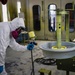 A worker in full protective gear is spray painting a large circular component, likely a vehicle wheel or rim, mounted on a rotating stand inside an industrial paint booth. The worker is wearing a white Tyvek suit, respirator mask, goggles, and blue nitrile gloves while operating a spray gun. The environment is a dedicated paint booth with yellow-coated walls and ventilation infrastructure.