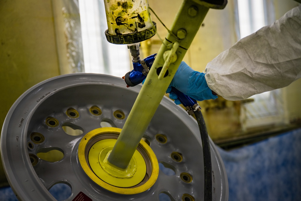 A worker in protective gear (white Tyvek suit and blue nitrile gloves) is operating a spray gun or dispensing tool over a large metal wheel or rim. The wheel appears to be mounted on a rotating fixture, and yellow material (likely paint, primer, or sealant) is being applied to the center hub area. This appears to be an industrial wheel refinishing or coating operation.