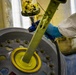 A worker in protective gear (white Tyvek suit and blue nitrile gloves) is operating a spray gun or dispensing tool over a large metal wheel or rim. The wheel appears to be mounted on a rotating fixture, and yellow material (likely paint, primer, or sealant) is being applied to the center hub area. This appears to be an industrial wheel refinishing or coating operation.