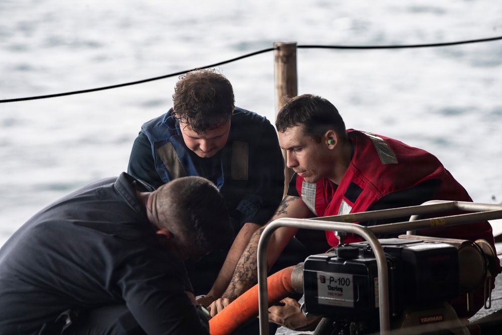 Three individuals are working together on what appears to be a naval or maritime vessel deck, focused on operating or maintaining a P-100 Darley pump. Two of the individuals are wearing safety vests — one red and one dark blue — suggesting a safety-conscious work environment. The scene takes place on open water, visible in the background.