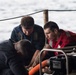 Three individuals are working together on what appears to be a naval or maritime vessel deck, focused on operating or maintaining a P-100 Darley pump. Two of the individuals are wearing safety vests — one red and one dark blue — suggesting a safety-conscious work environment. The scene takes place on open water, visible in the background.