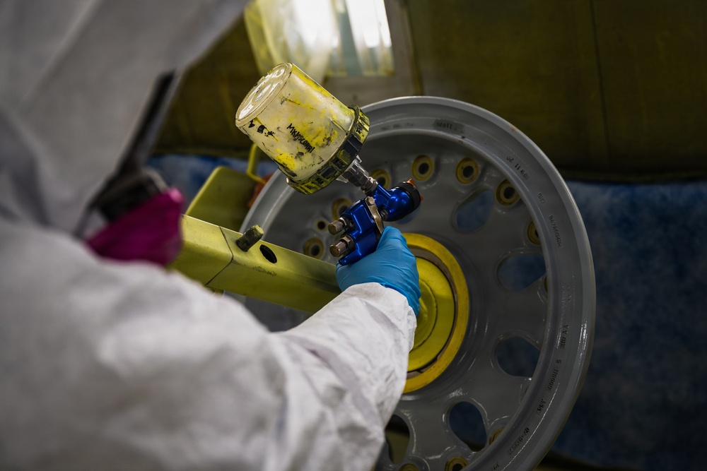 A technician in a white protective suit and blue nitrile gloves is using a pneumatic or power tool (blue-colored) on a large aircraft or aerospace wheel/hub assembly. The wheel rim is gray with a yellow center hub, featuring multiple bolt holes around its circumference. The work appears to be taking place in an industrial maintenance or manufacturing facility.
