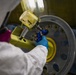 A technician in a white protective suit and blue nitrile gloves is using a pneumatic or power tool (blue-colored) on a large aircraft or aerospace wheel/hub assembly. The wheel rim is gray with a yellow center hub, featuring multiple bolt holes around its circumference. The work appears to be taking place in an industrial maintenance or manufacturing facility.