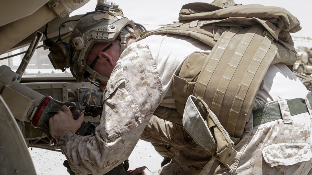A military service member wearing desert MARPAT camouflage uniform and tactical gear is leaning into a vehicle, appearing to perform maintenance or inspection work. The individual is equipped with a combat helmet with communications gear, body armor with MOLLE webbing, and a tactical backpack. The arid, bright environment suggests a desert operational setting.