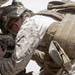 A military service member wearing desert MARPAT camouflage uniform and tactical gear is leaning into a vehicle, appearing to perform maintenance or inspection work. The individual is equipped with a combat helmet with communications gear, body armor with MOLLE webbing, and a tactical backpack. The arid, bright environment suggests a desert operational setting.