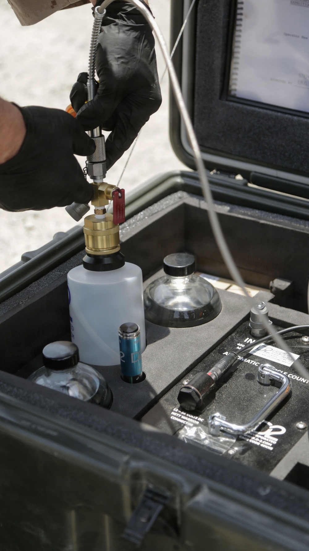 A person wearing black tactical gloves is operating a portable field testing kit housed in a ruggedized hard case. They are connecting a hose fitting to a brass valve assembly mounted on top of a white sample bottle. The kit appears to be a military or industrial-grade automatic particle counter used for fluid contamination analysis.