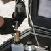 A person wearing black tactical gloves is operating a portable field testing kit housed in a ruggedized hard case. They are connecting a hose fitting to a brass valve assembly mounted on top of a white sample bottle. The kit appears to be a military or industrial-grade automatic particle counter used for fluid contamination analysis.