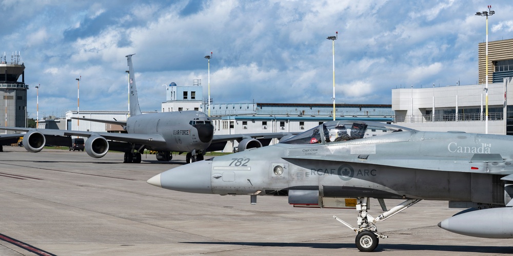The image shows a Royal Canadian Air Force (RCAF) CF-18 Hornet fighter jet (tail number 782) taxiing on an airport ramp, with a US Air Force KC-135 Stratotanker visible in the background. The scene appears to be at a joint military or civil-military airfield, with airport terminal buildings and a control tower visible. A pilot is seated in the cockpit of the Canadian fighter jet.