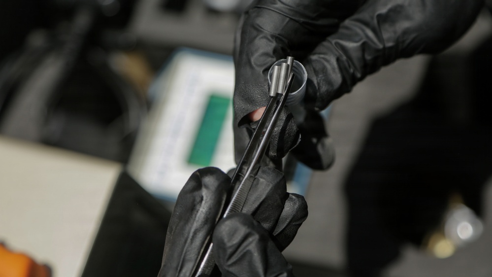A person wearing black nitrile/latex gloves is handling a dark metallic tool or instrument, likely in a technical or forensic/mechanical context. The item appears to be a slender metal rod or precision instrument being examined or manipulated. The background shows a blurred workspace with what appears to be documents or materials on a surface.