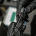 A person wearing black nitrile/latex gloves is handling a dark metallic tool or instrument, likely in a technical or forensic/mechanical context. The item appears to be a slender metal rod or precision instrument being examined or manipulated. The background shows a blurred workspace with what appears to be documents or materials on a surface.