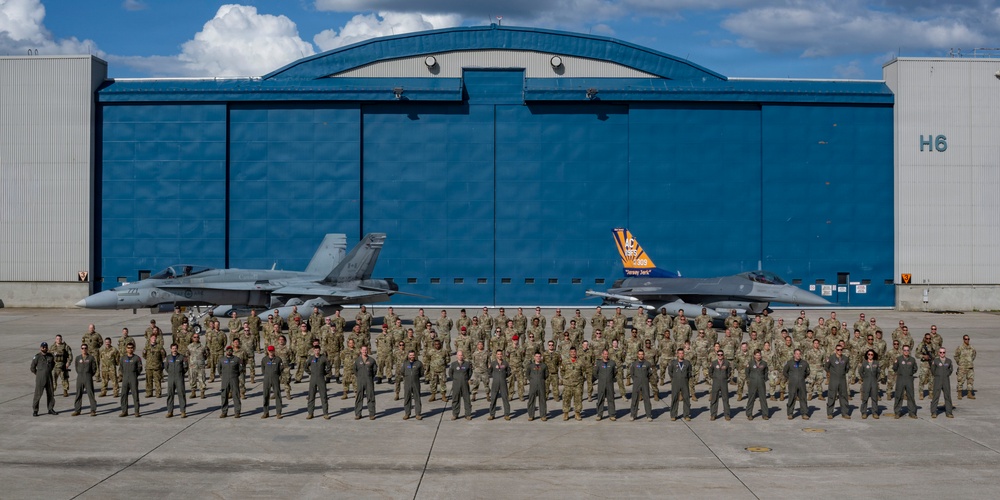 A large group of military personnel pose for a formal group photograph in front of a blue aircraft hangar labeled 'H6'. Two military jet aircraft, appearing to be F/A-18 Hornets or similar fighter jets, are positioned behind the personnel. One aircraft features a distinctive orange and blue commemorative tail marking, possibly indicating an anniversary or special designation.