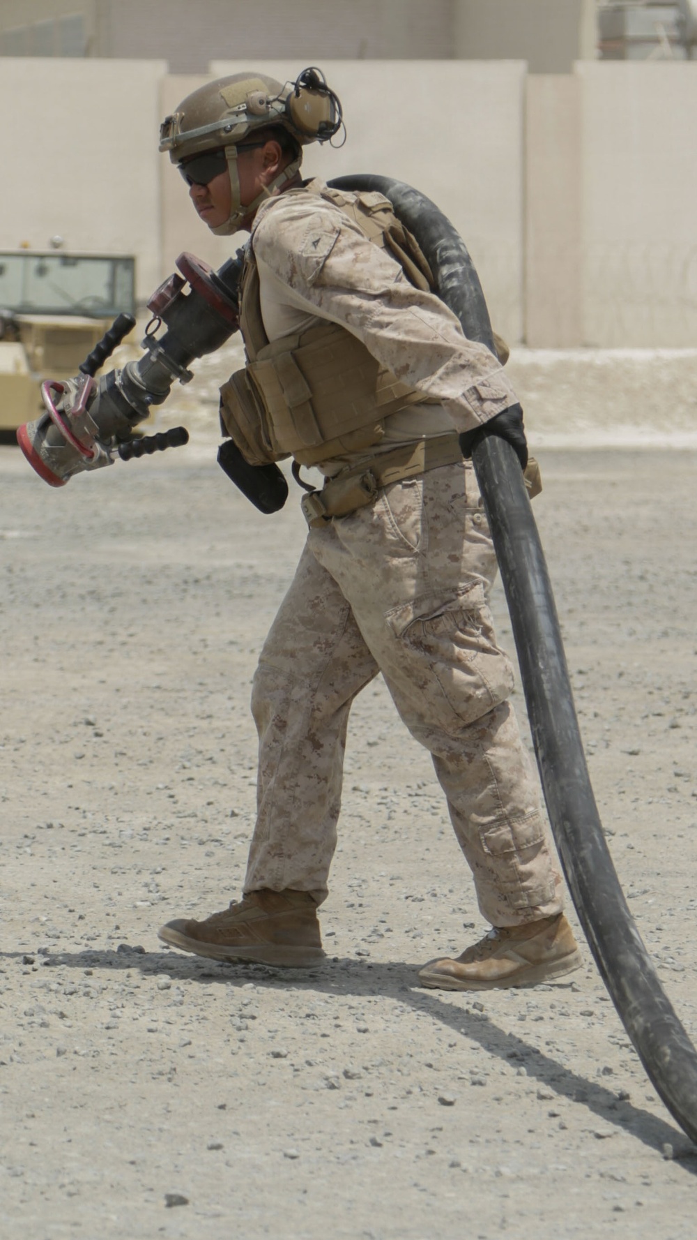 A military service member in desert camouflage (MARPAT pattern) is carrying a large black hose and a nozzle/coupling assembly across an arid, gravel-covered surface. The individual is wearing full combat gear including a ballistic helmet with communications equipment, body armor, and tactical pouches. The setting appears to be a forward operating base or military installation in a desert environment.