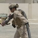 A military service member in desert camouflage (MARPAT pattern) is carrying a large black hose and a nozzle/coupling assembly across an arid, gravel-covered surface. The individual is wearing full combat gear including a ballistic helmet with communications equipment, body armor, and tactical pouches. The setting appears to be a forward operating base or military installation in a desert environment.
