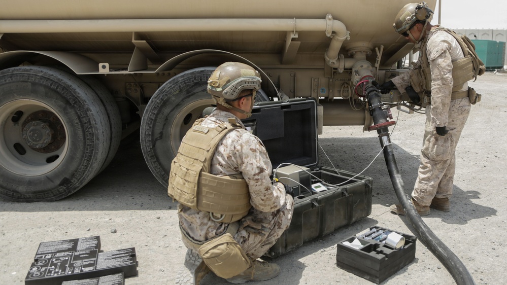 Two military personnel in desert camouflage uniforms are conducting what appears to be a fuel quality testing or sampling operation next to a large tan military tanker truck. One soldier is kneeling and operating a portable electronic equipment case, while the other is standing and handling a fuel hose connected to the tanker. The setting appears to be an arid, outdoor military installation or forward operating base.