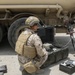 Two military personnel in desert camouflage uniforms are conducting what appears to be a fuel quality testing or sampling operation next to a large tan military tanker truck. One soldier is kneeling and operating a portable electronic equipment case, while the other is standing and handling a fuel hose connected to the tanker. The setting appears to be an arid, outdoor military installation or forward operating base.