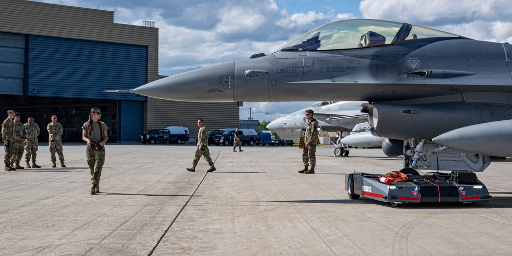 Military personnel in camouflage uniforms are gathered on an airfield tarmac alongside an F-16 Fighting Falcon jet fighter. A ground support tow vehicle is positioned under the aircraft's nose, suggesting maintenance or repositioning operations. The scene appears to take place at a military air base with hangars visible in the background.