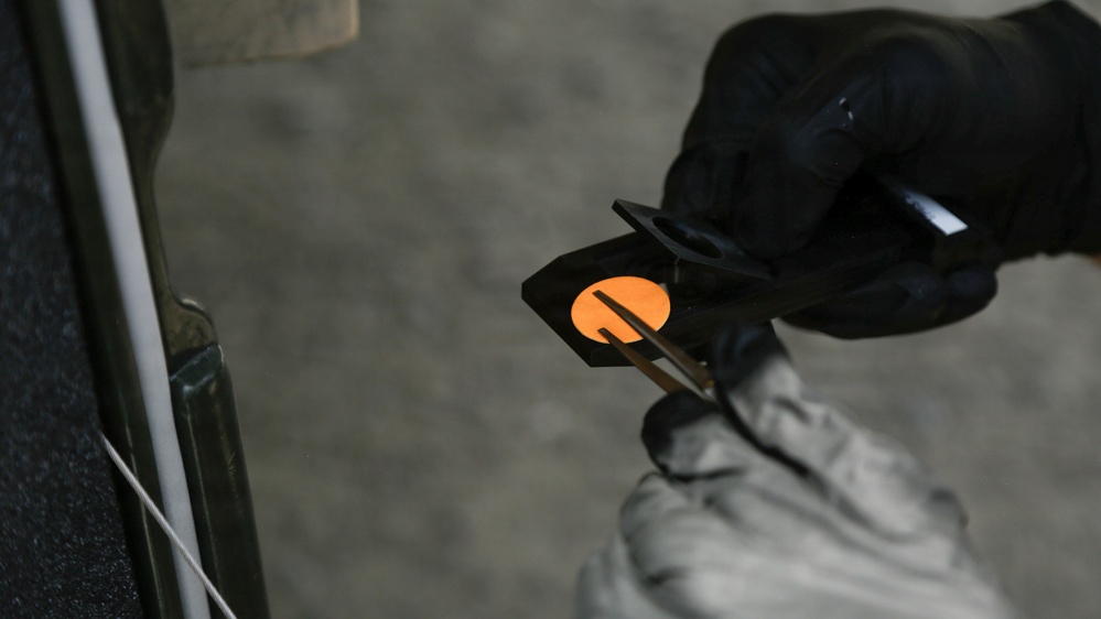A person wearing black gloves is holding a small black device or tool that contains a circular orange/amber-colored element, possibly a lens, filter, or indicator light. The item appears to be positioned near a vehicle door or panel edge visible on the left side of the frame. The close-up perspective suggests a technical or maintenance activity.