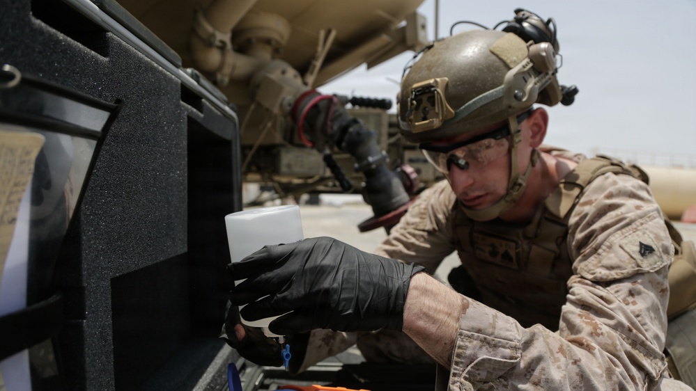 A military service member wearing desert camouflage (MARPAT pattern) and a tactical helmet is performing maintenance or inspection work on a military vehicle. The individual is wearing black nitrile gloves and appears to be working with a small white container near a vehicle compartment. The setting appears to be an arid, outdoor environment consistent with a forward operating base or motor pool.