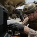 A military service member wearing desert camouflage (MARPAT pattern) and a tactical helmet is performing maintenance or inspection work on a military vehicle. The individual is wearing black nitrile gloves and appears to be working with a small white container near a vehicle compartment. The setting appears to be an arid, outdoor environment consistent with a forward operating base or motor pool.