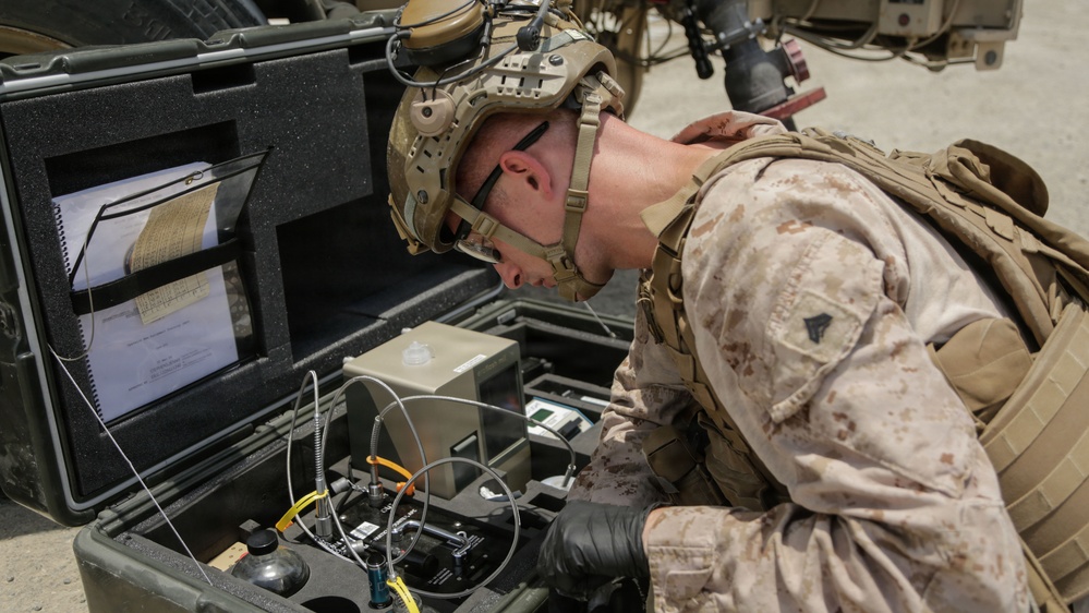 A U.S. Marine in full combat gear, including a tactical helmet and MARPAT desert camouflage uniform, is closely examining a ruggedized equipment case containing electronic or communications hardware. The case appears to be a portable field system with cables, connectors, and mounted components, along with reference documents in the lid. The setting appears to be an outdoor operational environment near a military vehicle.