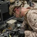 A U.S. Marine in full combat gear, including a tactical helmet and MARPAT desert camouflage uniform, is closely examining a ruggedized equipment case containing electronic or communications hardware. The case appears to be a portable field system with cables, connectors, and mounted components, along with reference documents in the lid. The setting appears to be an outdoor operational environment near a military vehicle.