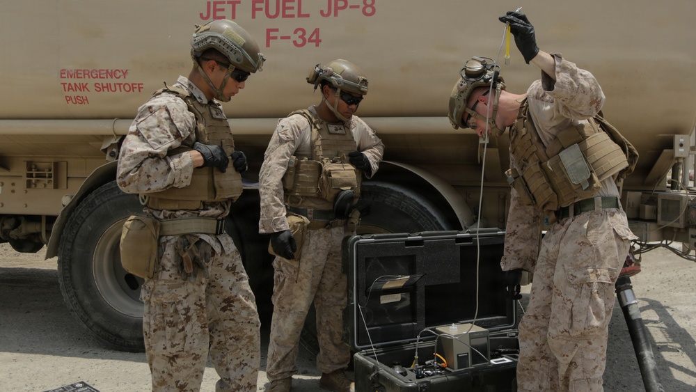Three military personnel in desert camouflage uniforms are working near a large fuel tanker labeled 'Jet Fuel JP-8 F-34'. They appear to be conducting fuel quality testing or sampling operations using a portable equipment case with cables and instruments. The setting appears to be an arid, outdoor military installation or forward operating base.