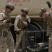 Three military personnel in desert camouflage uniforms are working near a large fuel tanker labeled 'Jet Fuel JP-8 F-34'. They appear to be conducting fuel quality testing or sampling operations using a portable equipment case with cables and instruments. The setting appears to be an arid, outdoor military installation or forward operating base.