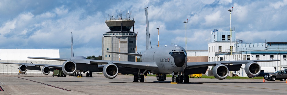 A large U.S. Air Force military tanker aircraft, identifiable as a KC-135 Stratotanker, is parked on the flight line of a military air base. The aircraft is positioned facing the camera with four jet engines visible beneath its swept wings. Behind the aircraft stands an air traffic control tower and associated base infrastructure buildings.