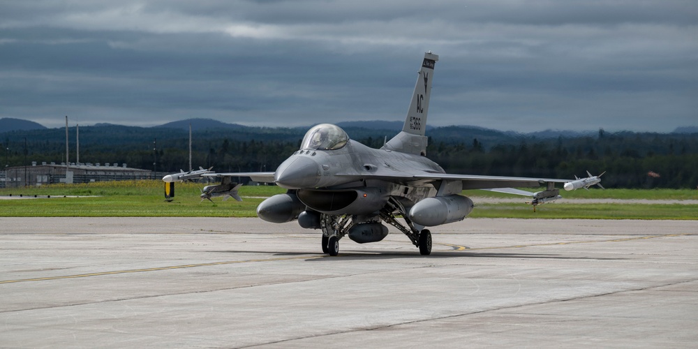 A U.S. Air Force F-16 Fighting Falcon jet fighter is taxiing on a concrete runway apron at a military airbase. The aircraft bears 'AC' tail codes, indicating it belongs to the Atlantic City Air National Guard (177th Fighter Wing). The scene is set under overcast skies with forested hills and base infrastructure visible in the background.