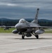 A U.S. Air Force F-16 Fighting Falcon jet fighter is taxiing on a concrete runway apron at a military airbase. The aircraft bears 'AC' tail codes, indicating it belongs to the Atlantic City Air National Guard (177th Fighter Wing). The scene is set under overcast skies with forested hills and base infrastructure visible in the background.