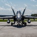 A front-facing view of what appears to be an F/A-18 Hornet or Super Hornet jet fighter parked on a concrete tarmac at a military airbase. A ground crew member in military fatigues is crouched beneath the aircraft's nose, likely performing a pre-flight inspection or maintenance check. The aircraft is in a gray paint scheme with twin vertical stabilizers and wingtip fuel tanks visible.