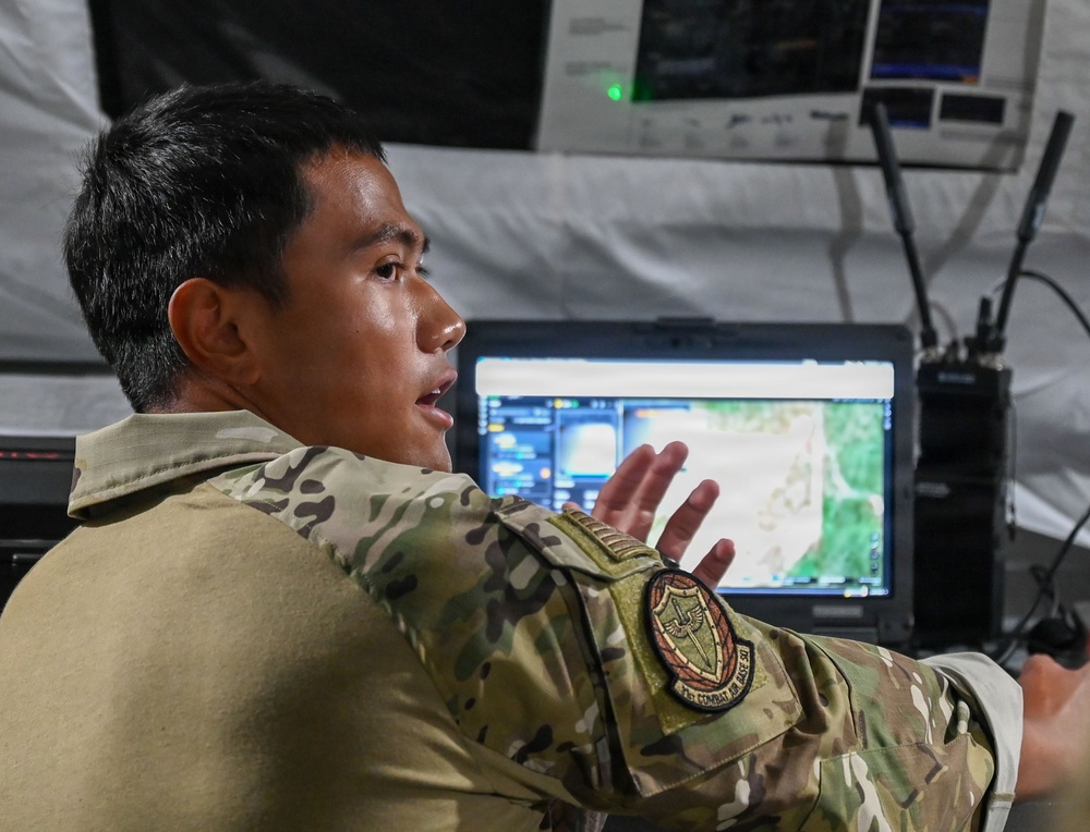 A military personnel member in multicam/OCP camouflage uniform is seated at a tactical operations workstation inside what appears to be a military tent or command post. The individual is speaking and gesturing with their hands, likely briefing or explaining information displayed on a monitor showing a map or aerial imagery. A tactical radio and additional screens are visible in the background.