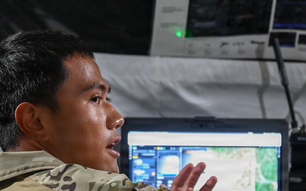 A military personnel member in multicam/OCP camouflage uniform is seated at a tactical operations workstation inside what appears to be a military tent or command post. The individual is speaking and gesturing with their hands, likely briefing or explaining information displayed on a monitor showing a map or aerial imagery. A tactical radio and additional screens are visible in the background.