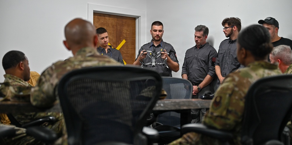 A briefing or demonstration session is taking place in a room where civilian instructors are presenting a small drone/quadcopter to a group of military personnel in camouflage uniforms. The central figure, dressed in a gray shirt, is holding and displaying the drone while others observe attentively. The setting appears to be a training or classroom environment.