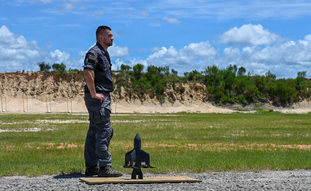 A man in military-style gray tactical clothing stands outdoors on a gravel surface next to what appears to be a small model rocket or munition replica placed on a wooden board. The setting appears to be an open training range or test field with a sandy berm, green grass, and trees in the background under a partly cloudy sky. The individual appears to be observing or overseeing some kind of outdoor activity or demonstration.