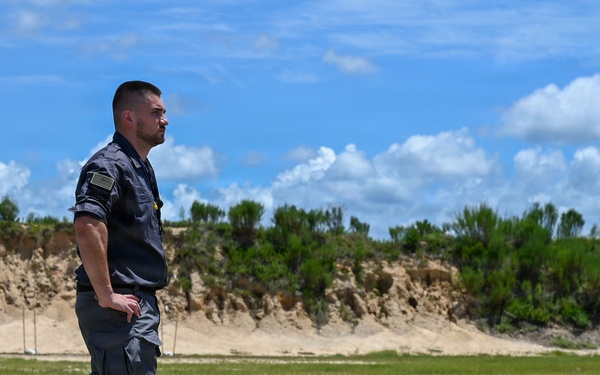 A man in military-style gray tactical clothing stands outdoors on a gravel surface next to what appears to be a small model rocket or munition replica placed on a wooden board. The setting appears to be an open training range or test field with a sandy berm, green grass, and trees in the background under a partly cloudy sky. The individual appears to be observing or overseeing some kind of outdoor activity or demonstration.
