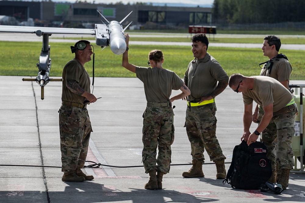 Five military personnel in OCP (Operational Camouflage Pattern) uniforms are gathered on an airfield tarmac near the wing of a military aircraft. One individual is holding up what appears to be an air-to-air missile mounted under the aircraft wing, while the group appears to be engaged in discussion or instruction. The setting appears to be a NATO or US Air Force base based on the infrastructure visible in the background.