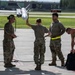 Five military personnel in OCP (Operational Camouflage Pattern) uniforms are gathered on an airfield tarmac near the wing of a military aircraft. One individual is holding up what appears to be an air-to-air missile mounted under the aircraft wing, while the group appears to be engaged in discussion or instruction. The setting appears to be a NATO or US Air Force base based on the infrastructure visible in the background.