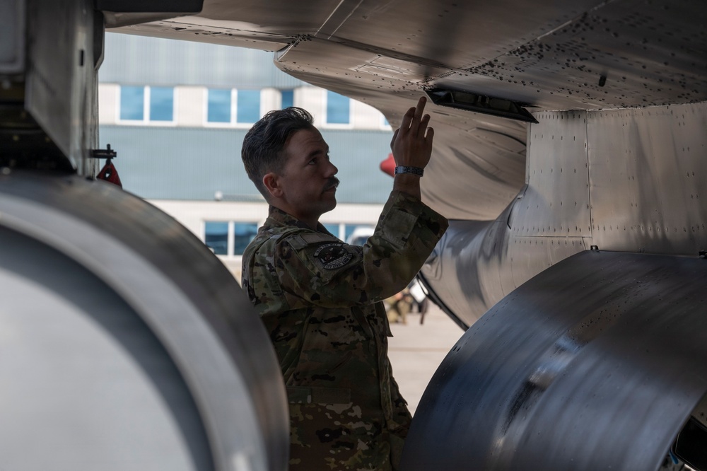 A military personnel member in camouflage uniform is conducting a pre-flight or maintenance inspection on the underside of a military aircraft. The individual is closely examining or touching a panel or component beneath the aircraft's wing or fuselage. The setting appears to be an outdoor flight line or tarmac near a building.