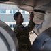 A military personnel member in camouflage uniform is conducting a pre-flight or maintenance inspection on the underside of a military aircraft. The individual is closely examining or touching a panel or component beneath the aircraft's wing or fuselage. The setting appears to be an outdoor flight line or tarmac near a building.