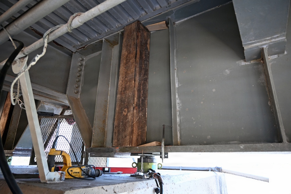 The image shows an industrial or construction setting, likely underneath a bridge or heavy steel structure, where repair or maintenance work appears to be in progress. A large wooden timber beam is positioned vertically between steel structural members, possibly being used as a temporary support or shoring element. Tools and equipment are visible on a concrete surface below.