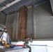 The image shows an industrial or construction setting, likely underneath a bridge or heavy steel structure, where repair or maintenance work appears to be in progress. A large wooden timber beam is positioned vertically between steel structural members, possibly being used as a temporary support or shoring element. Tools and equipment are visible on a concrete surface below.