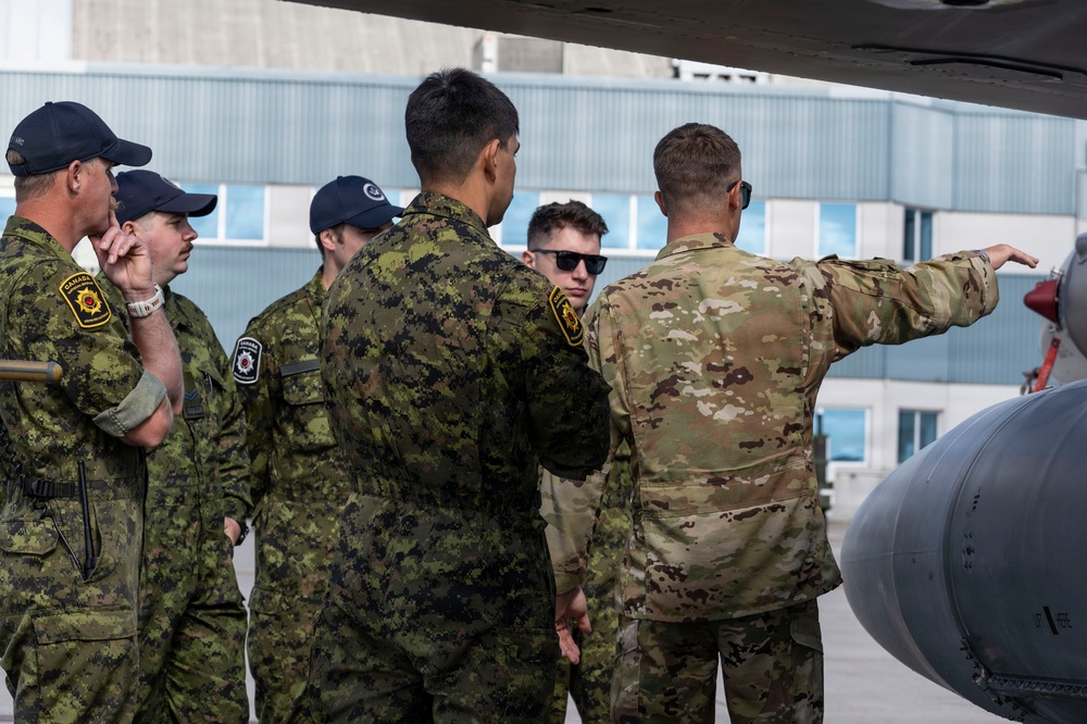 A group of military personnel in two distinct uniforms are gathered on an airfield tarmac near what appears to be an aircraft. Several individuals wearing Canadian Armed Forces CADPAT (Canadian Disruptive Pattern) uniforms with 'CANADA' shoulder patches are being briefed by a person in US-style OCP (Operational Camouflage Pattern) uniform. The person in OCP is pointing toward a component of the aircraft, suggesting a technical briefing or familiarization tour.