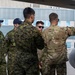A group of military personnel in two distinct uniforms are gathered on an airfield tarmac near what appears to be an aircraft. Several individuals wearing Canadian Armed Forces CADPAT (Canadian Disruptive Pattern) uniforms with 'CANADA' shoulder patches are being briefed by a person in US-style OCP (Operational Camouflage Pattern) uniform. The person in OCP is pointing toward a component of the aircraft, suggesting a technical briefing or familiarization tour.