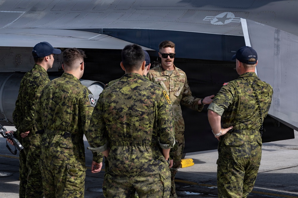 A U.S. Air Force service member in MultiCam/OCP uniform is briefing or conducting a walkthrough with a group of Canadian Armed Forces personnel next to a military aircraft, likely an F-35 or similar stealth jet. The U.S. airman, wearing sunglasses, appears to be explaining features of the aircraft while the Canadian personnel listen attentively. The setting appears to be an outdoor flight line or tarmac.